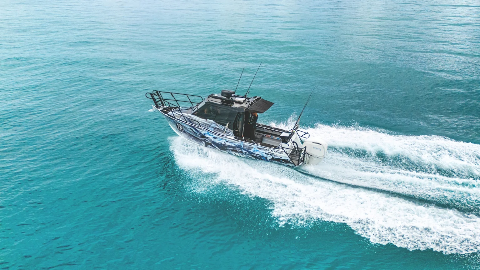 An overhead shot of a fishing boat with a blue and white design, branded "HUTCHWILCO 70" and "BOAT SHOW", speeds through turquoise water, leaving a white wake.