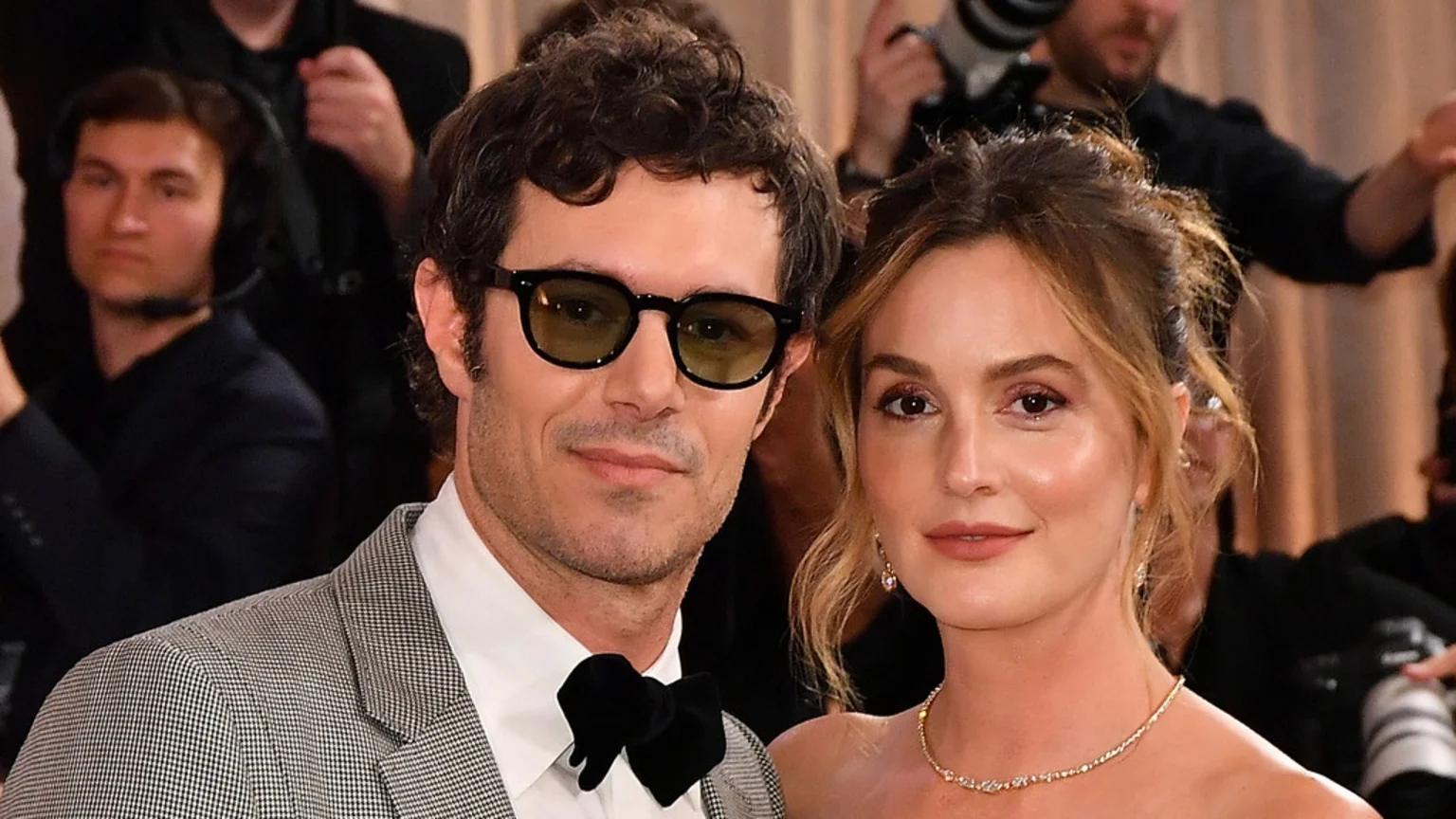 Adam Brody, in sunglasses and a plaid blazer, stands beside Leighton Meester, who wears a delicate diamond necklace, as they both look forward at a public event.