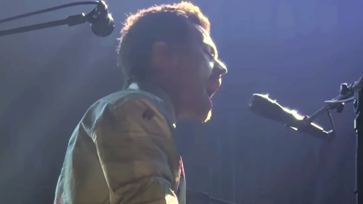 A person with curly hair, seen in profile, is singing or shouting into a microphone on a dimly lit stage, with bright backlighting highlighting their head.