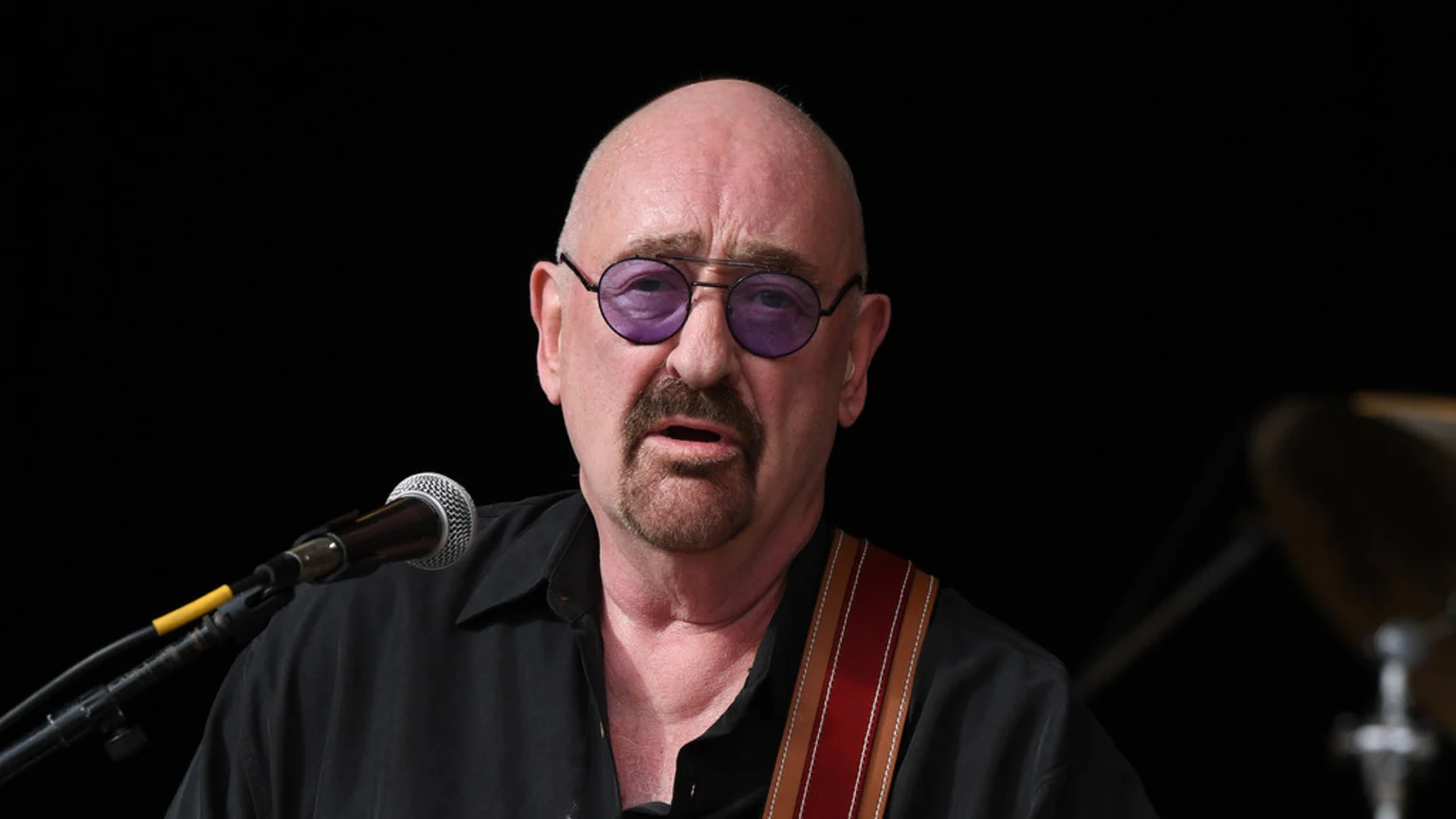 A bald man with a goatee and purple-tinted glasses, wearing a black shirt and a brown and red strap, speaks into a microphone against a dark background.