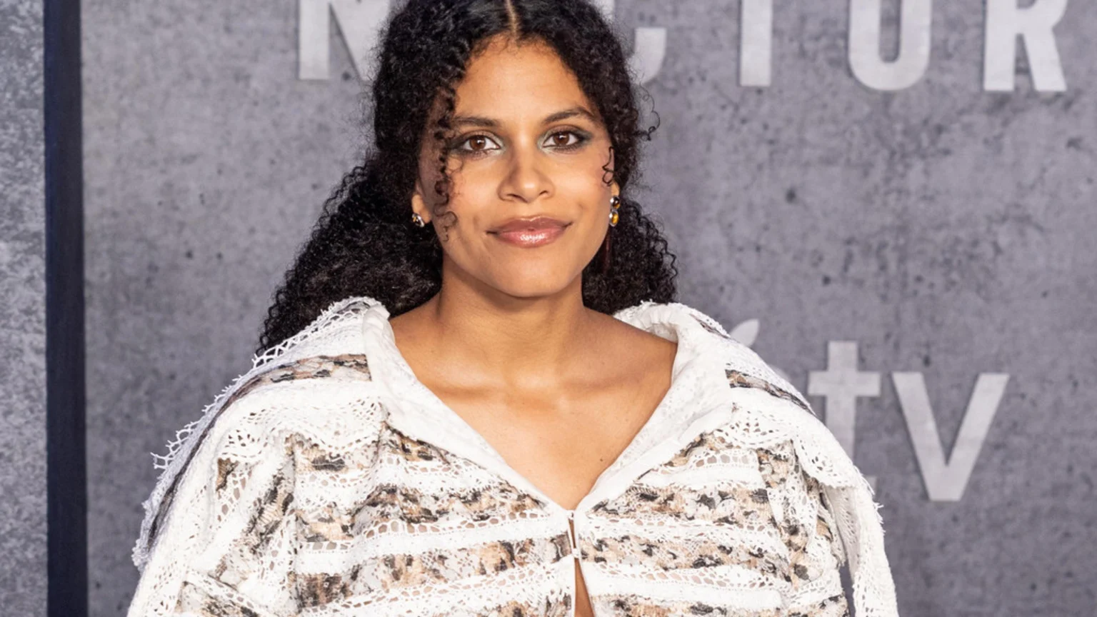 A woman with curly dark hair and light brown skin smiles subtly, wearing a white and brown patterned lace top against a grey background with white text.