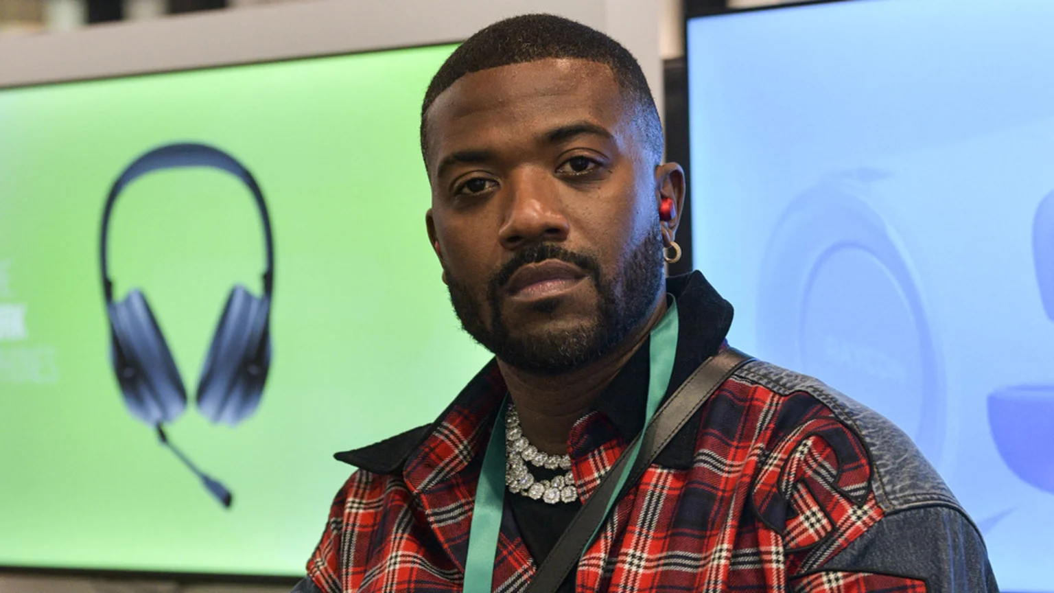 A man with a beard and short hair, wearing a red plaid shirt, diamond necklaces, a gold earring, and a red earbud, looks at the camera. Two screens behind him show headphones and a partially visible "RAYCON" product.