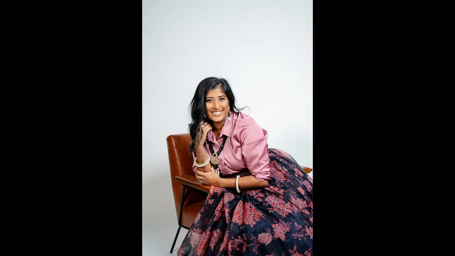 A smiling woman with dark hair sits in a brown armchair, wearing a pink long-sleeved shirt, a black floral skirt, and ornate jewelry.