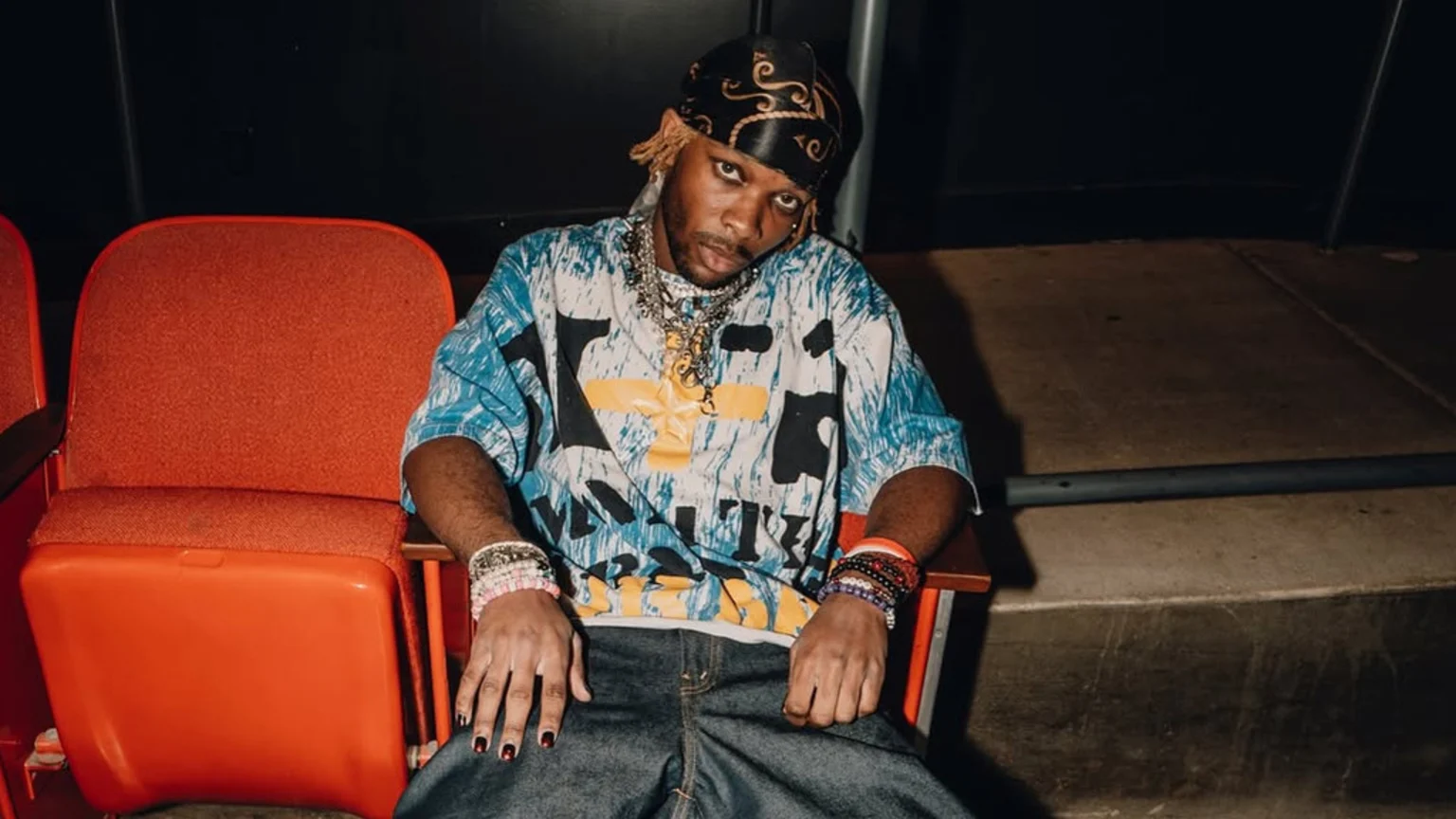 A person with a black patterned durag and light hair sits in an orange theater seat, wearing an oversized blue and white patterned t-shirt with a yellow cross design, multiple chains, bracelets, and dark nail polish.