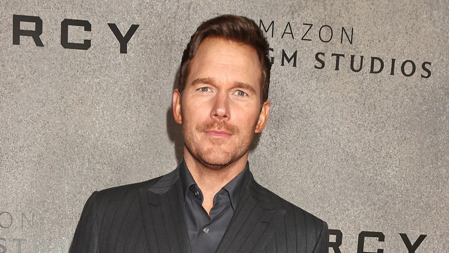 Actor Chris Pratt looks at the camera, sporting a beard and wearing a dark pinstripe suit, in front of a gray wall with visible text "MAZON GM STUDIOS".