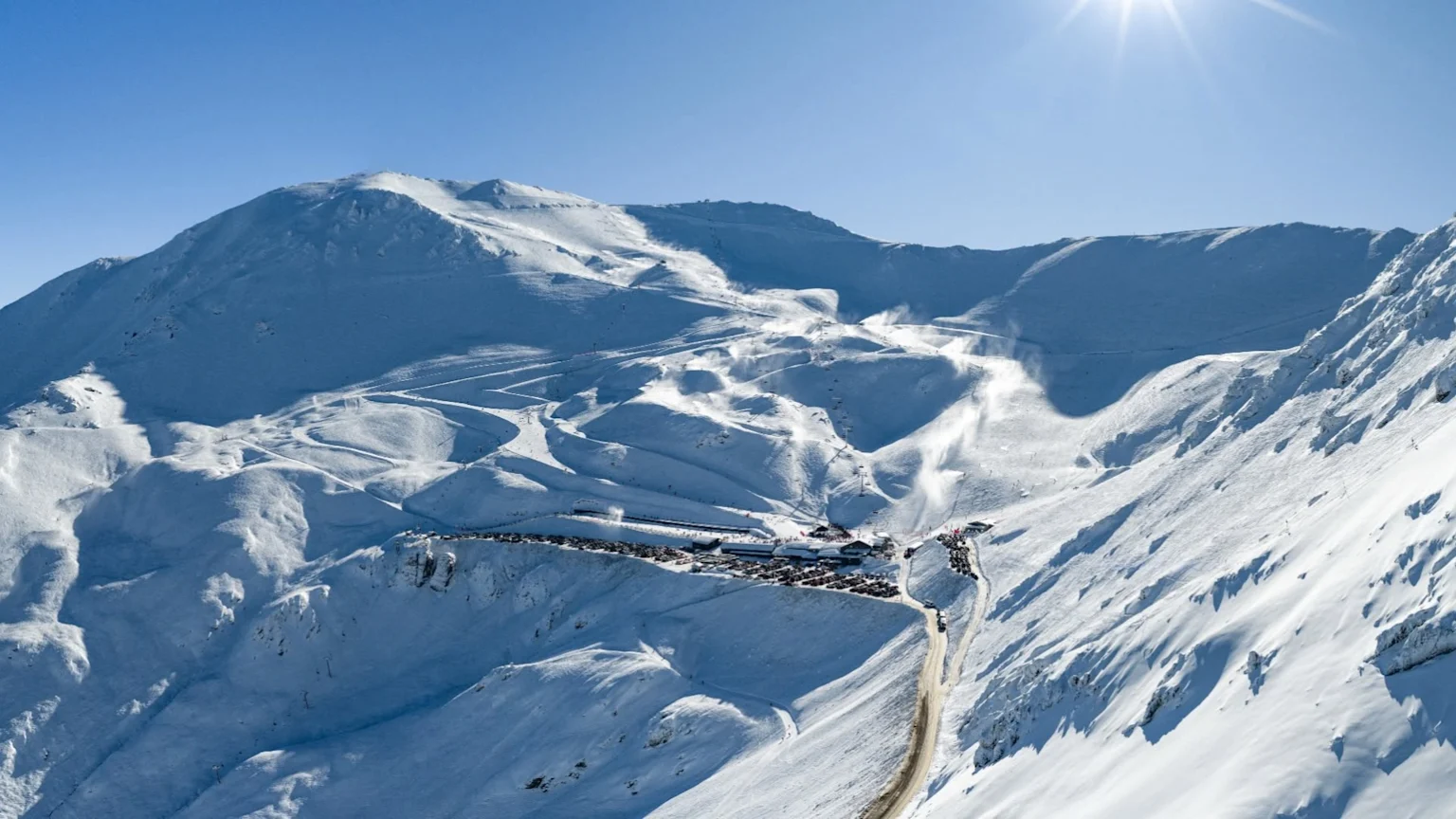 An aerial view of a busy snow-covered ski resort, showing numerous ski slopes, active chairlifts, a bustling base area with buildings and a full parking lot, all under a bright blue sky.
