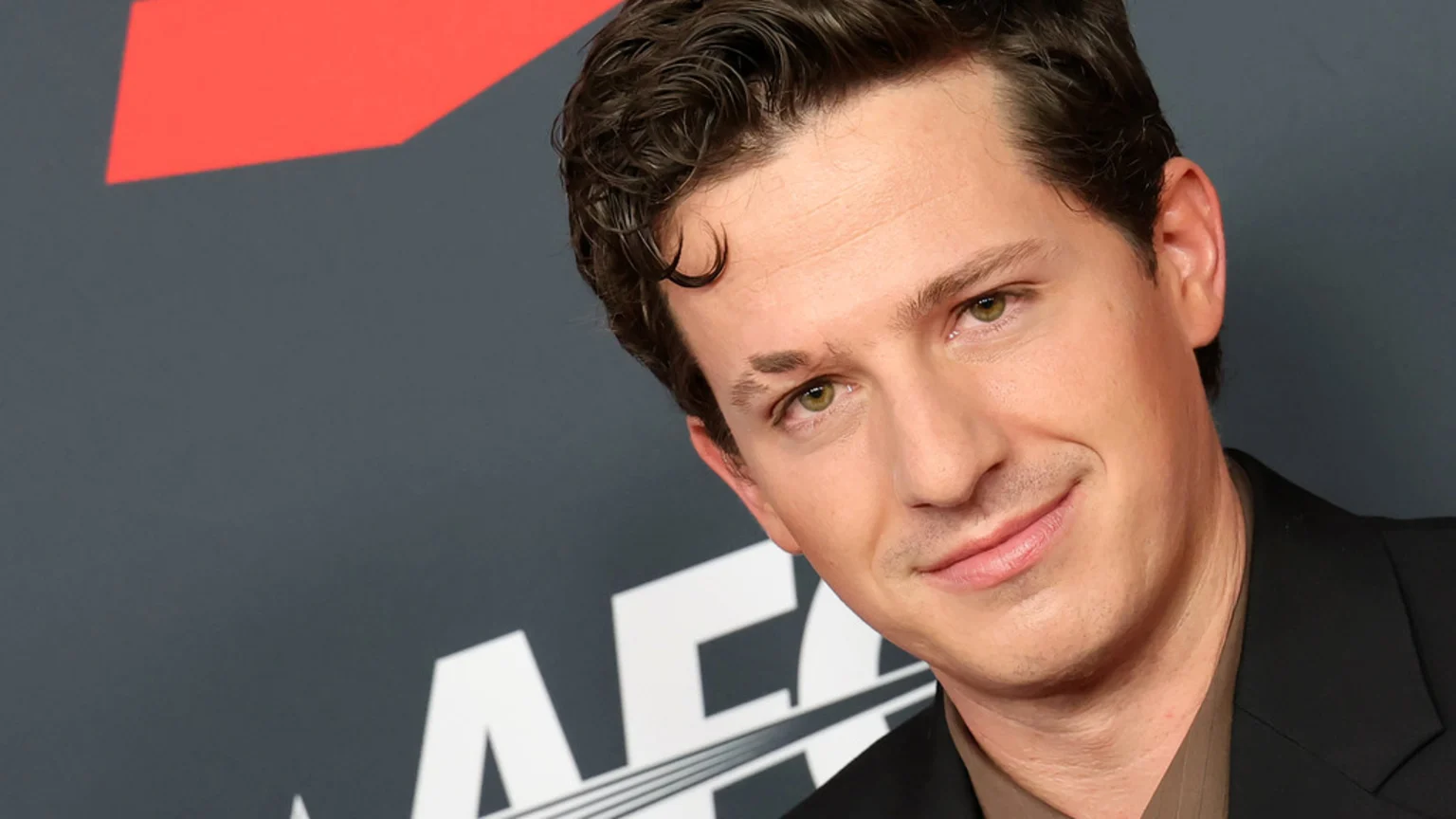 A close-up headshot of Charlie Puth smiling slightly, featuring his dark curly hair, green eyes, and distinct eyebrow scar, set against a dark gray background with red and white graphic elements.