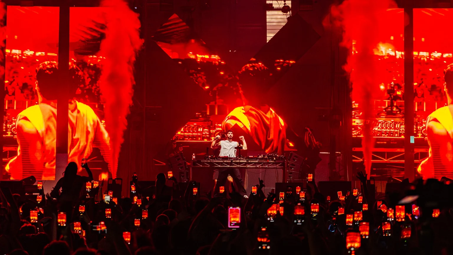 A DJ performs on a red-lit stage with large screens displaying red visuals and smoke plumes, while a crowd in the foreground holds up phones with glowing red lights.