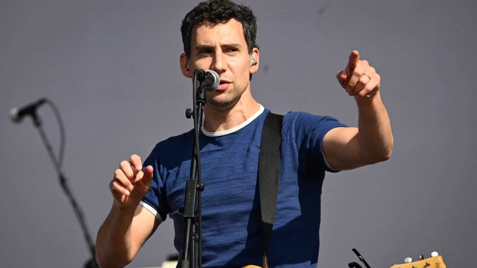 A man with dark, curly hair and a beard sings into a microphone, wearing a blue t-shirt and guitar strap, pointing his right index finger forward on a stage with a gray background.