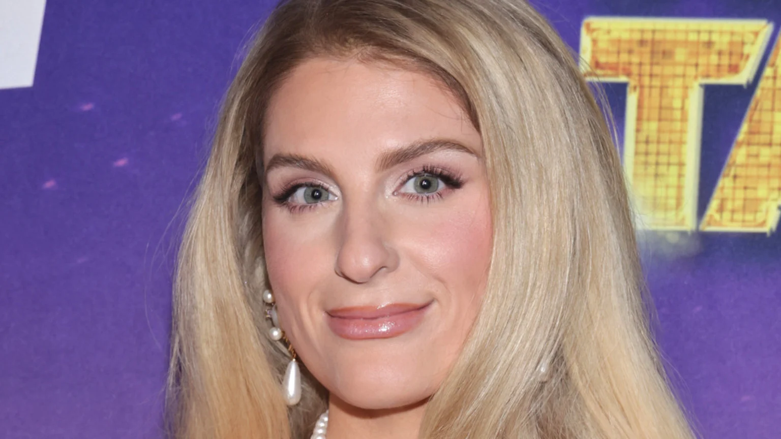 A close-up of singer Meghan Trainor with long blonde hair, light eyes, and a slight smile, wearing pearl earrings against a purple background.
