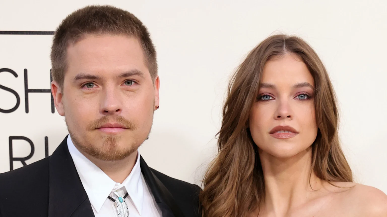 Dylan Sprouse, with a short haircut and beard, wearing a suit and bolo tie, poses beside Barbara Palvin, who has long brown wavy hair, wearing a strapless top and soft makeup.