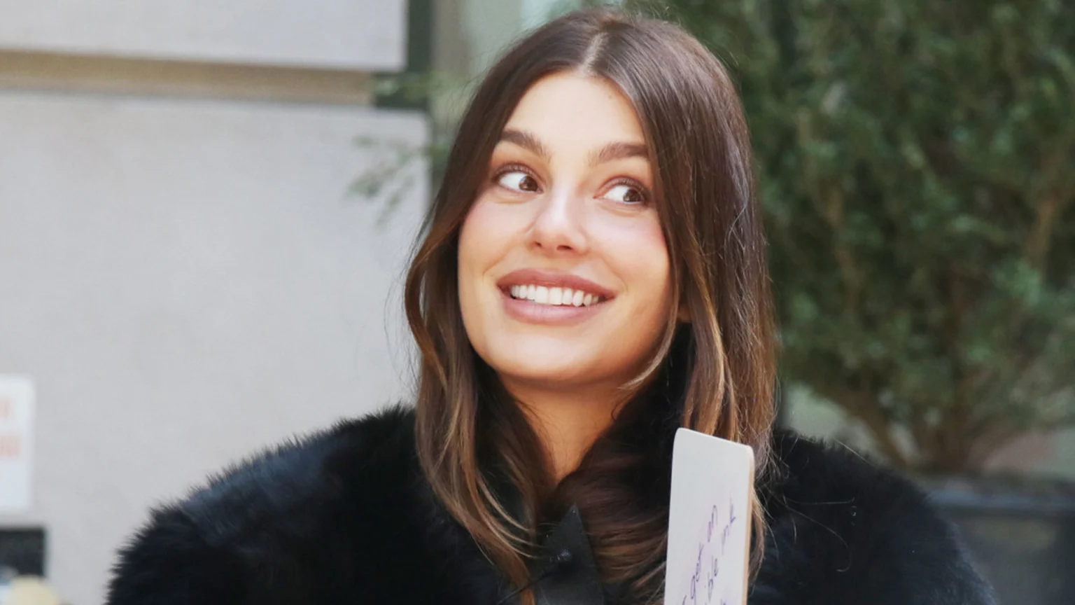 A smiling woman with long brown hair looks to the right, wearing a dark furry top and holding a white card, with a blurred background of a building and foliage.