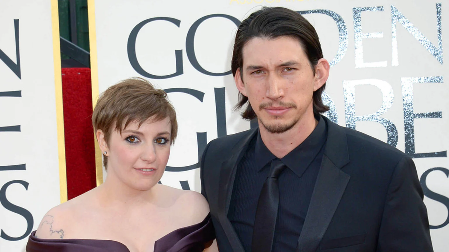 Lena Dunham in a dark off-the-shoulder dress and Adam Driver in a black suit pose at what appears to be the Golden Globe Awards.