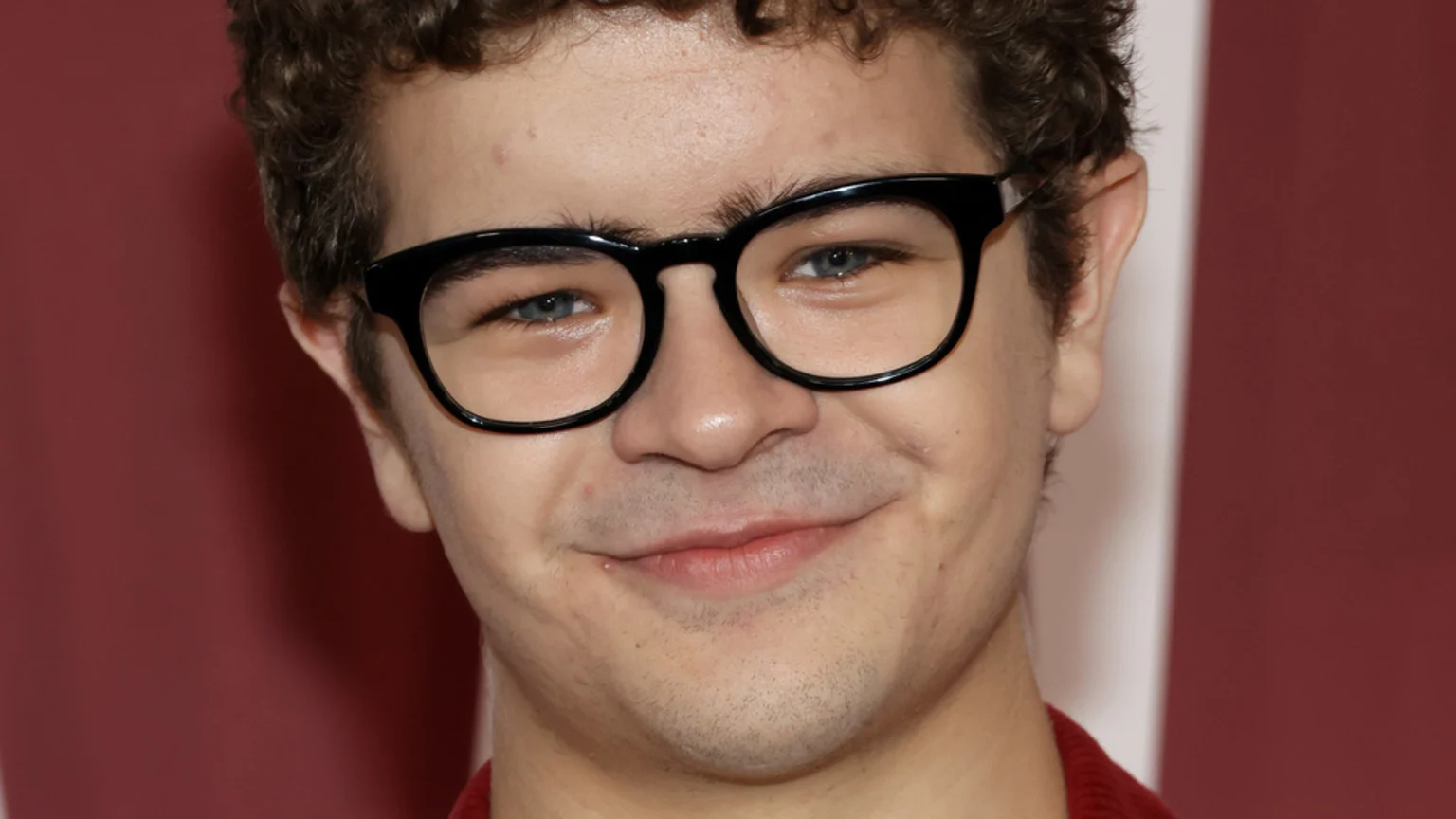Close-up of Gaten Matarazzo smiling, wearing black-rimmed glasses, with curly brown hair and blue eyes, against a blurred red background.