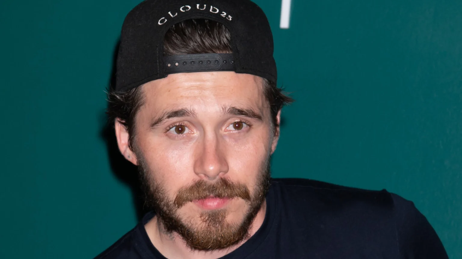 A man with a beard, wearing a black cap turned backward with "CLOUD23" on it, and a black t-shirt, looks directly at the camera against a teal background.