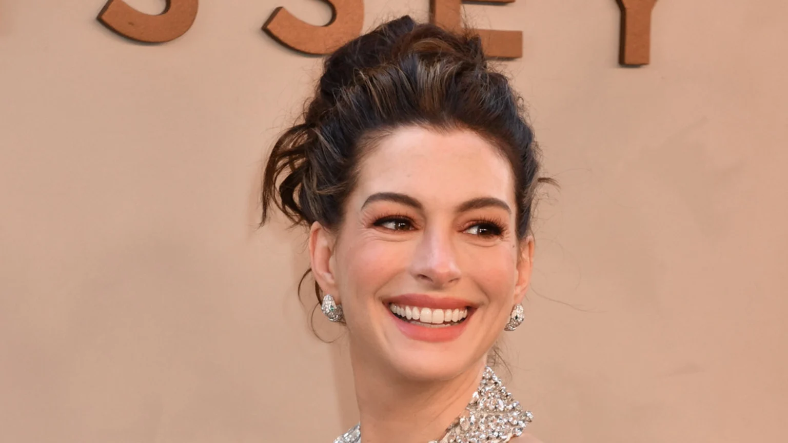 A close-up of Anne Hathaway smiling broadly, with her dark hair pulled back and wearing sparkly earrings and a top, against a light brown background with wooden letters.