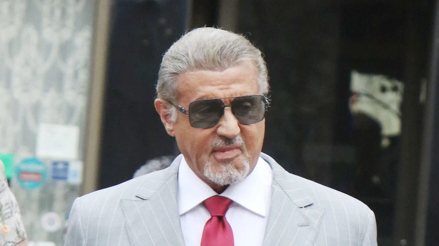 Sylvester Stallone, with grey hair and a goatee, wears sunglasses, a grey pinstripe suit, a white shirt, and a red tie.