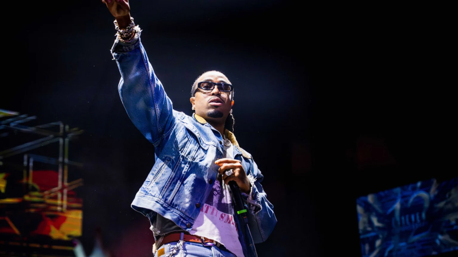 A man in a denim jacket and sunglasses raises his arm while performing on a dark stage with lights in the background.