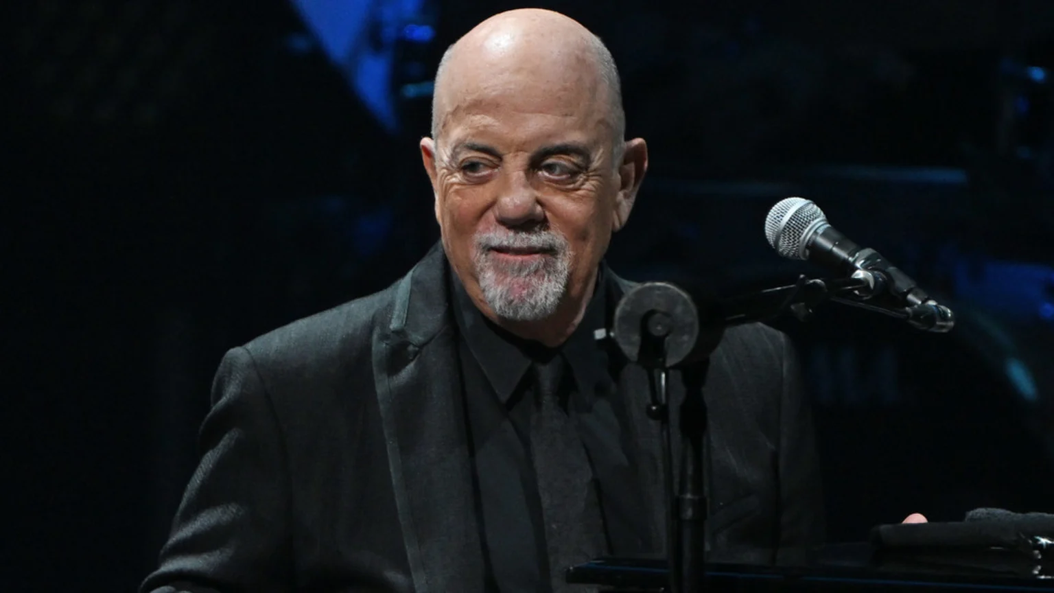 Billy Joel, a bald man with a grey goatee, smiles faintly while wearing a dark suit and tie, with a microphone positioned to his right against a dark background.