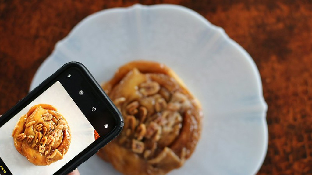 A hand holds a smartphone, taking a photo of a sticky bun topped with nuts and a glossy glaze on a white scalloped plate, with the phone's screen displaying the captured image.