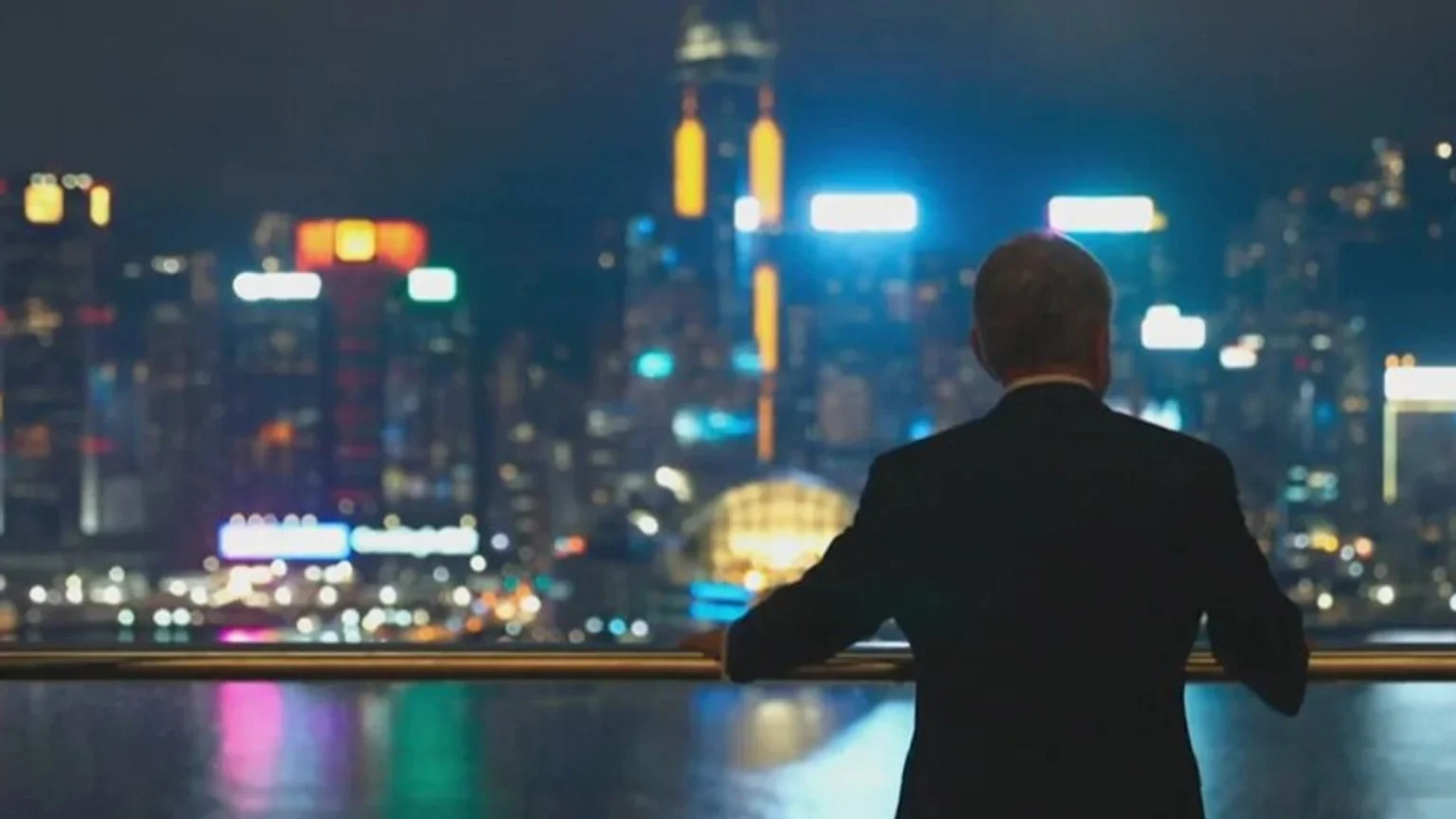 A man in a dark suit stands with his back to the camera, leaning on a railing and gazing at an out-of-focus, brightly lit city skyline at night, with colorful reflections on the water.