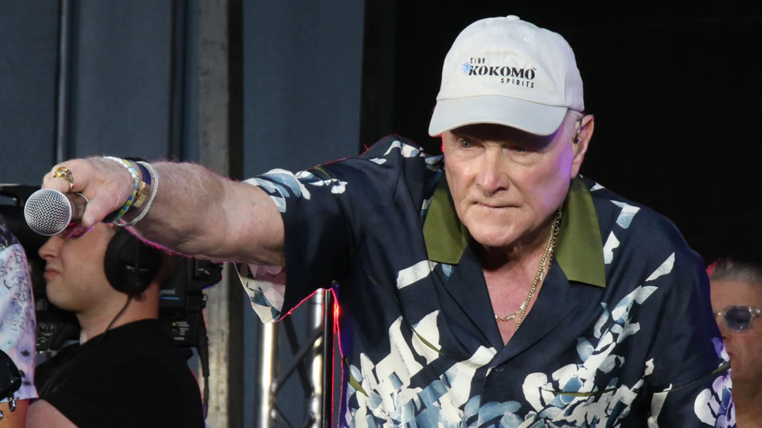 An older male performer in a white "KOKOMO SPIRITS" baseball cap and floral shirt holds a microphone, pointing left, with a camera person visible.