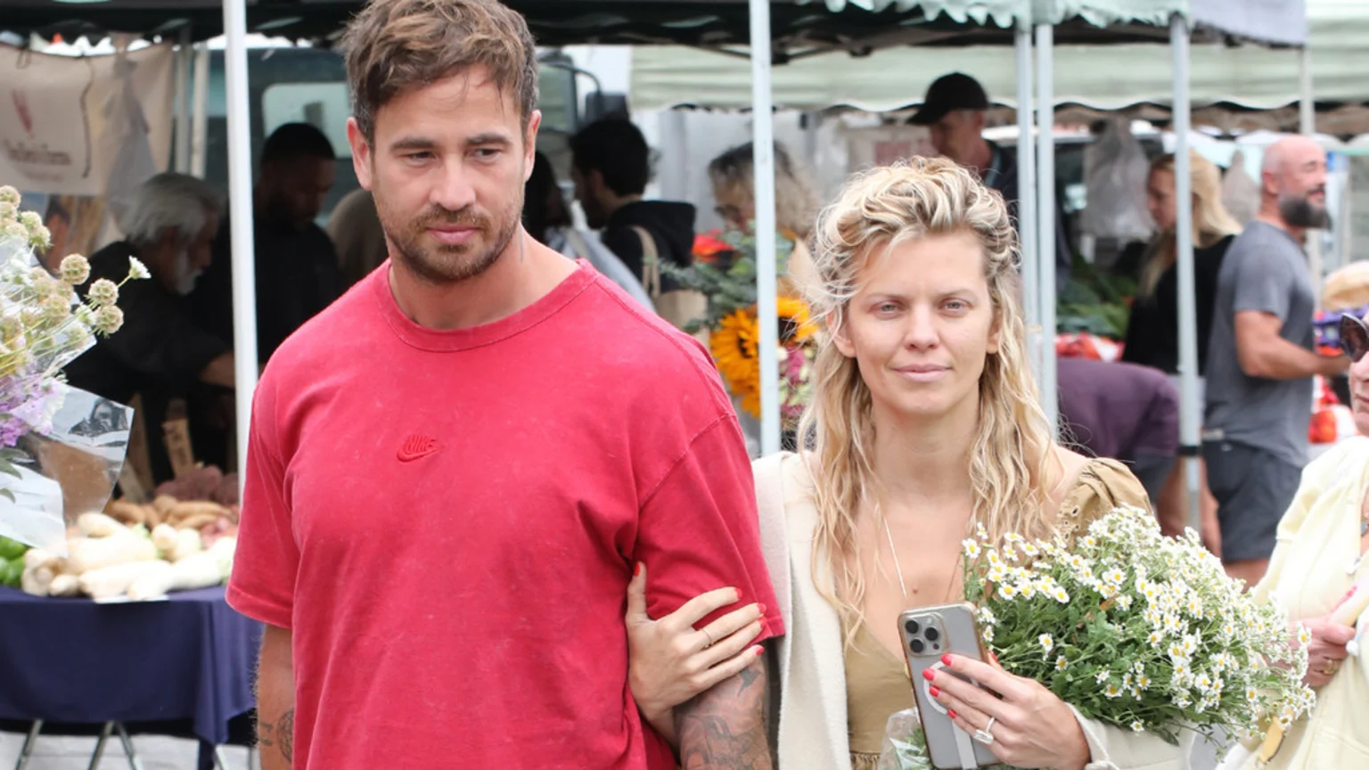 A man in a red shirt and a woman with blonde hair holding flowers walk through an outdoor market, with the woman holding the man's arm.