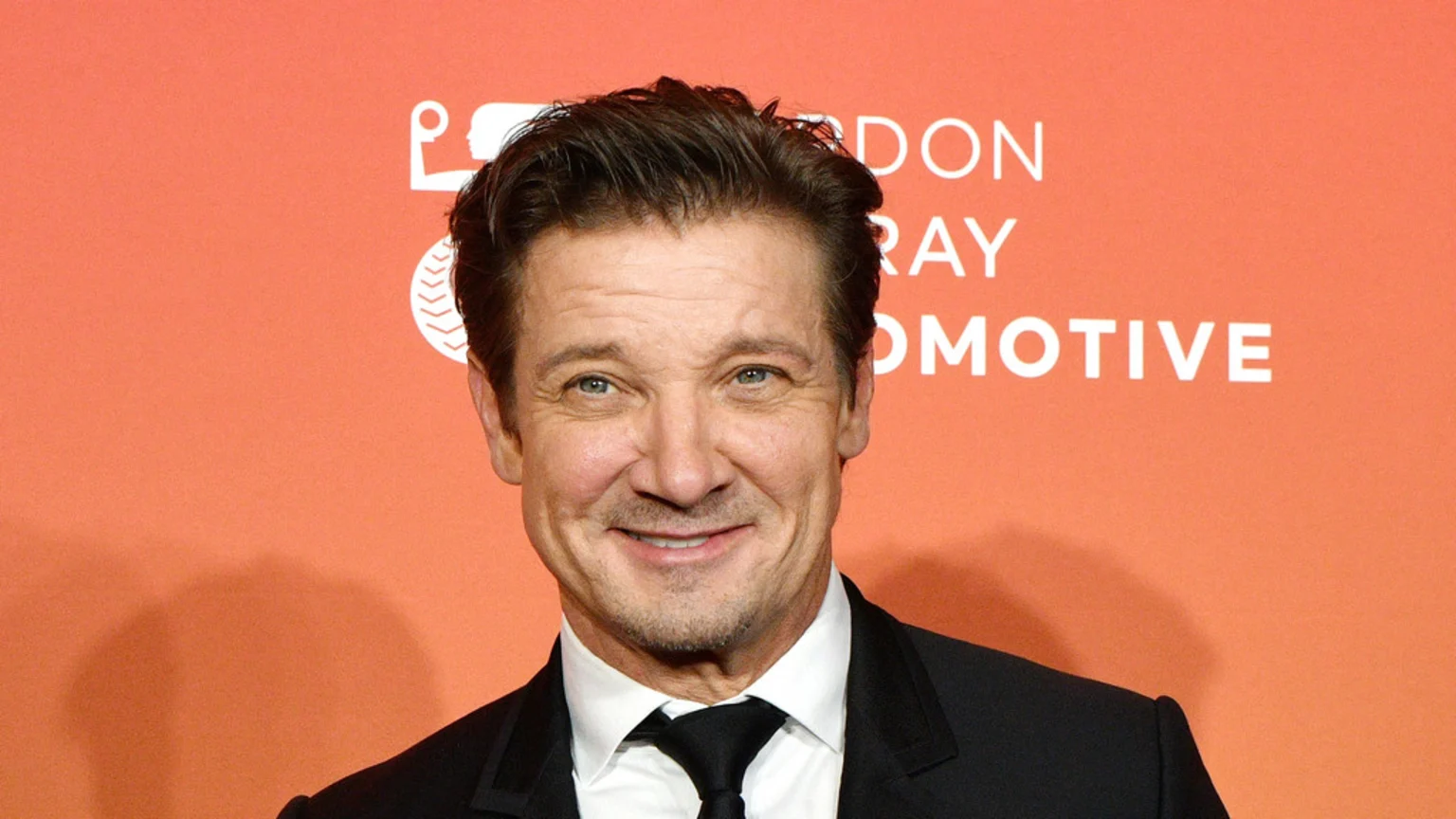 Jeremy Renner smiles, wearing a black suit and tie, against an orange background with partial white text visible.