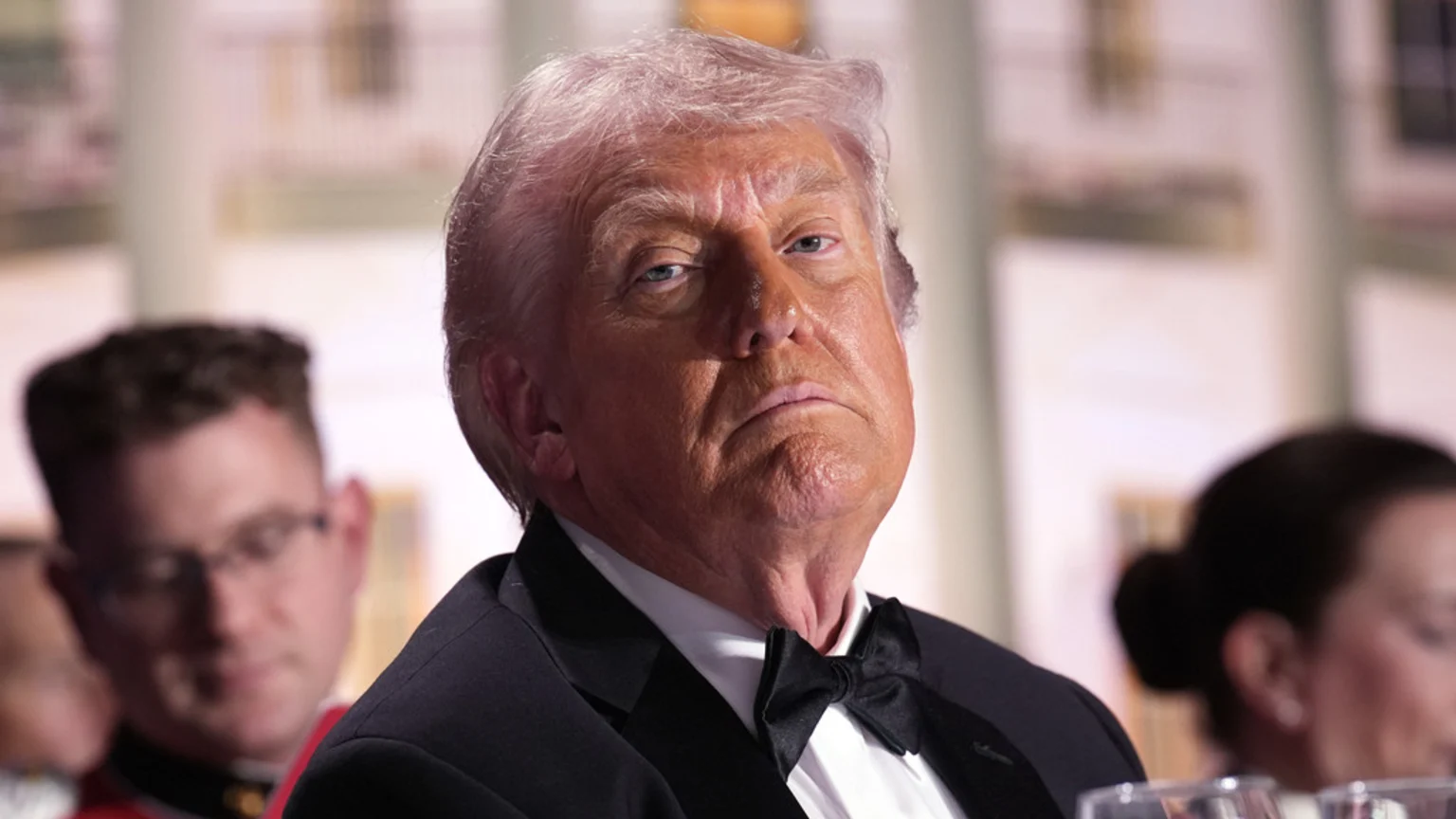 Donald Trump, wearing a tuxedo and bow tie, looks sternly with a tanned face and light hair, with blurred people and a building in the background.
