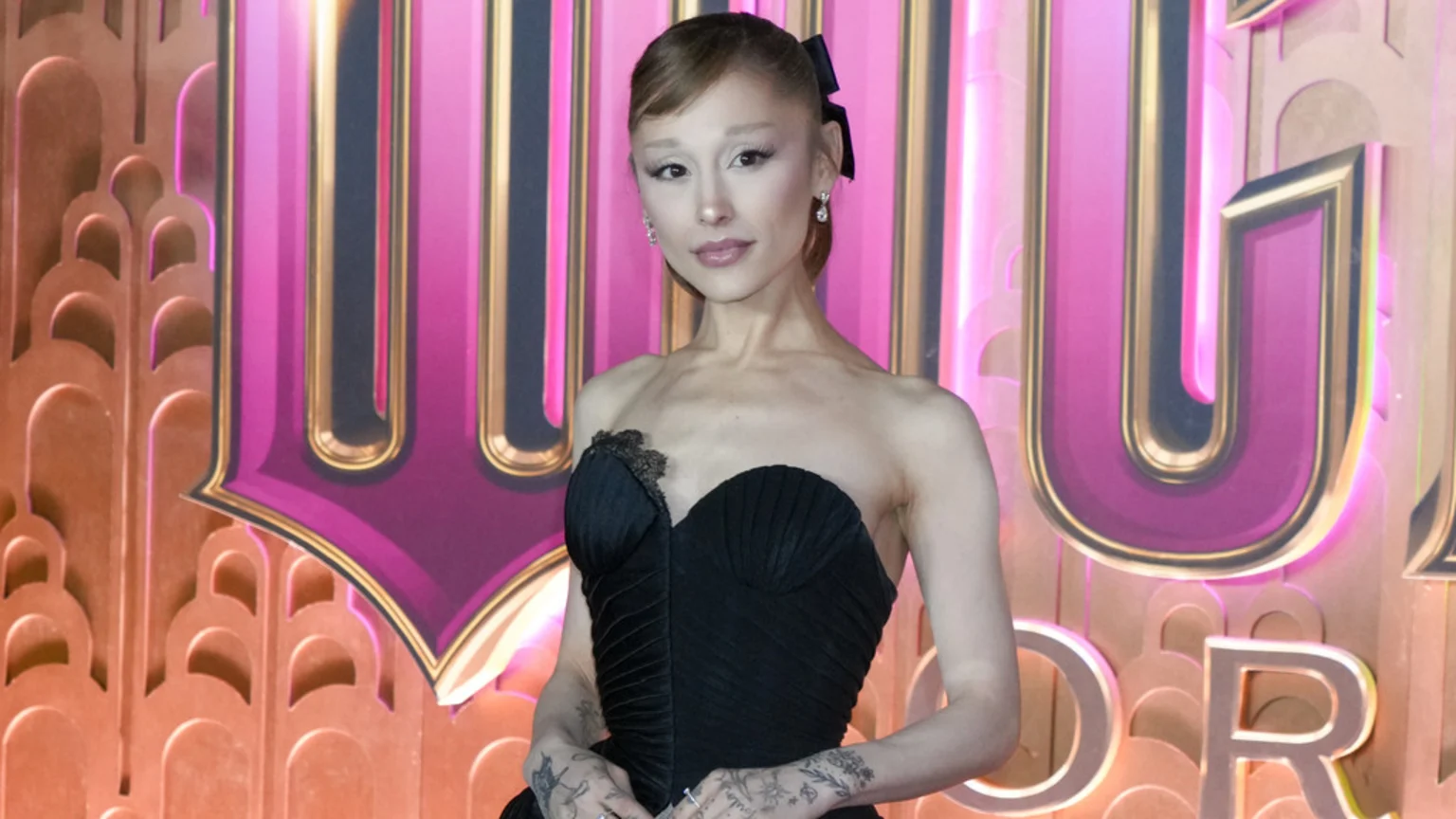 Ariana Grande smiles slightly, wearing a black strapless gown and a black bow in her hair, with a vibrant pink and gold art deco-themed background featuring large letters like "U" and "OR".