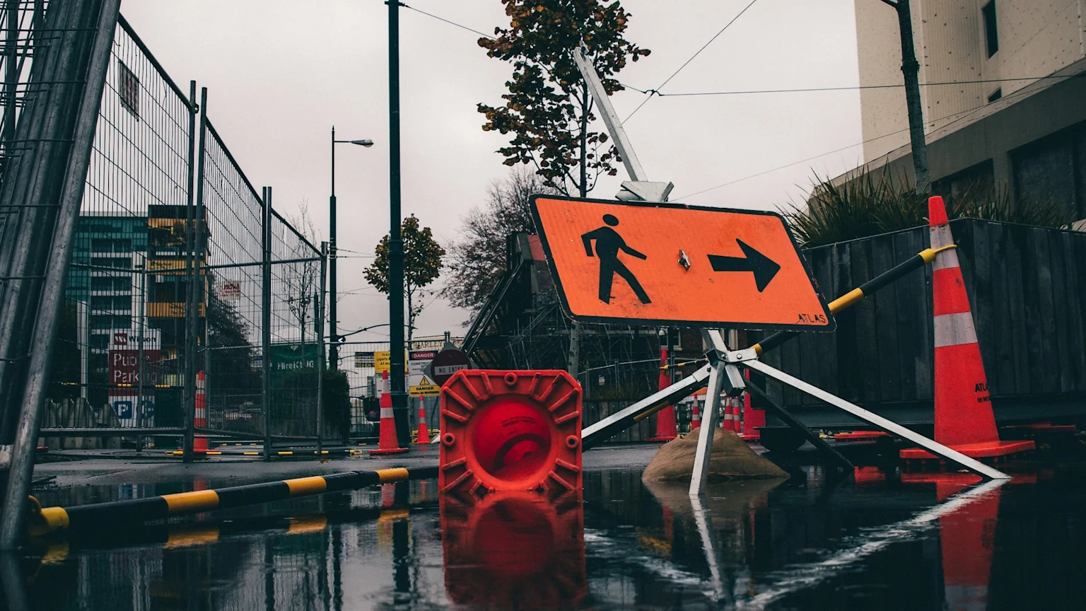 An orange pedestrian detour sign stands on a wet street with construction fences, traffic cones, and a fallen barrier, all reflected in puddles under an overcast sky.