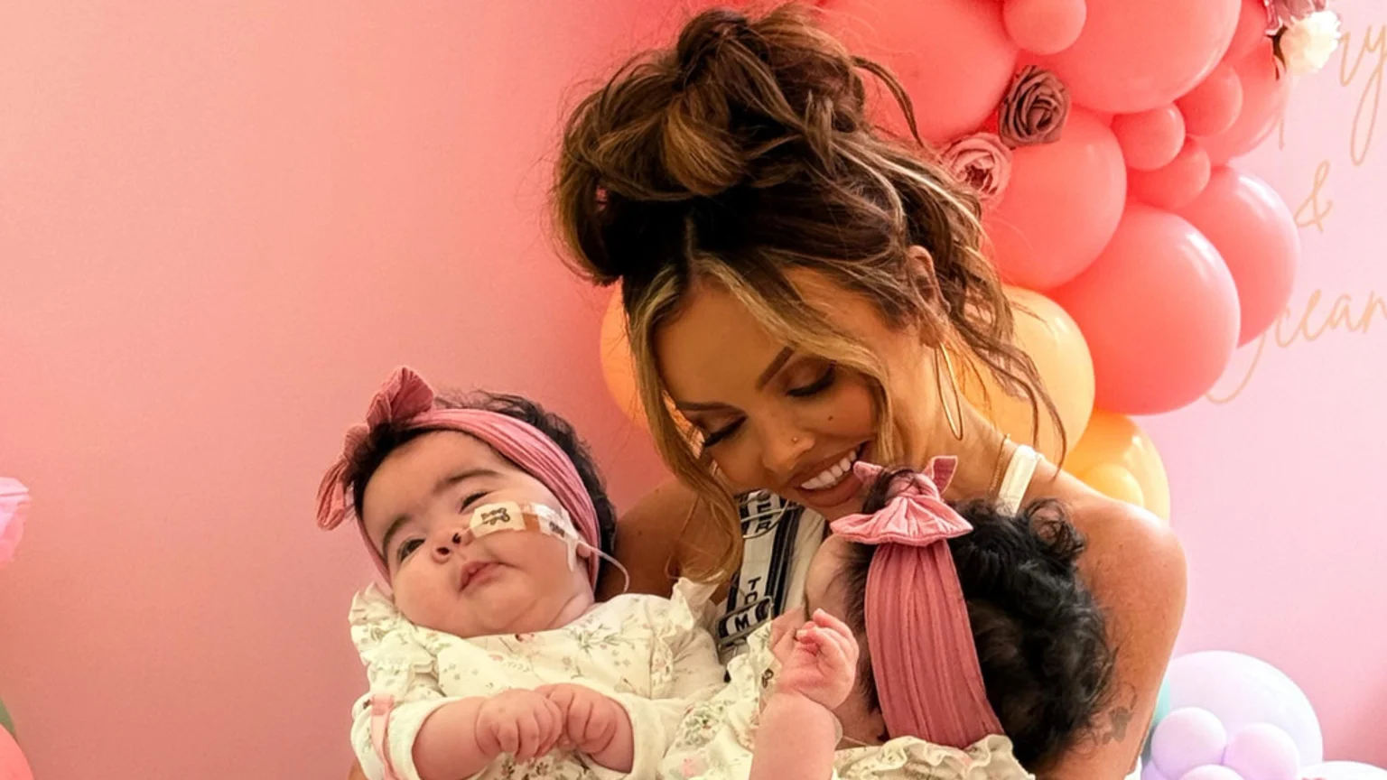 A smiling woman with a bun holds two babies in a pink room; one baby wears a pink headband and has a feeding tube taped to her nose.