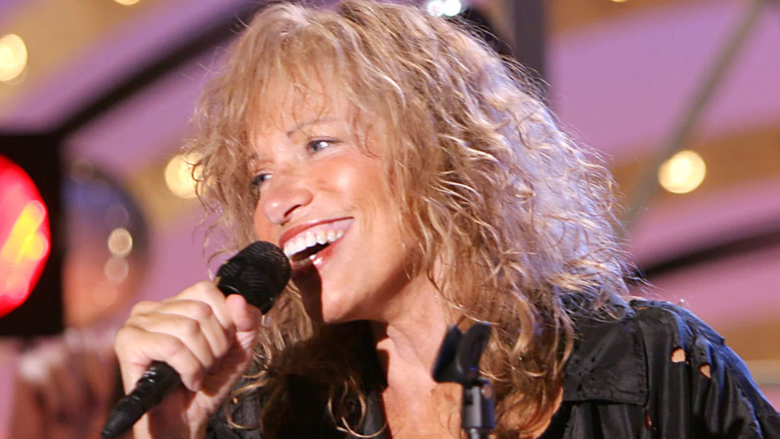 A smiling woman with long, curly blonde hair sings into a microphone, wearing a dark distressed shirt, with blurred lights in the background.