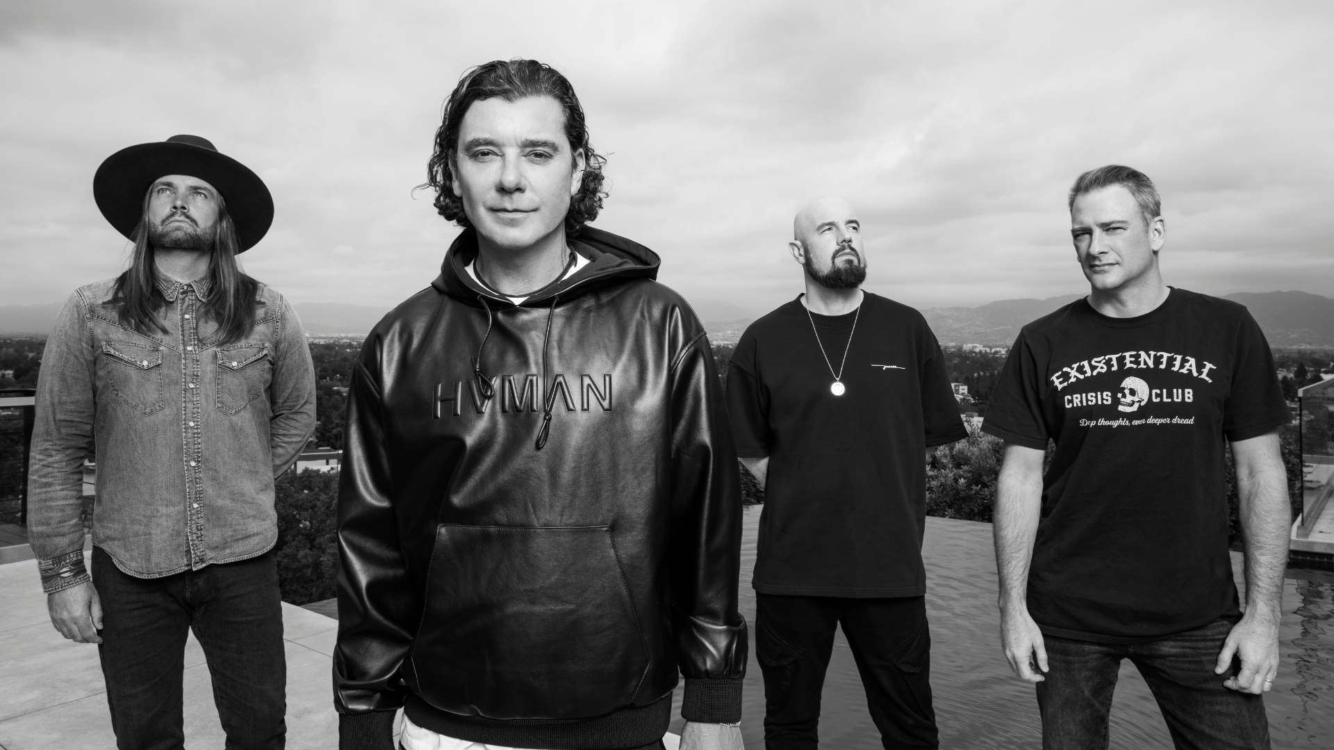 A black and white portrait of four men in casual attire standing outdoors; one wears a hat, another a 'HVMAN' hoodie, and a third an 'Existential Crisis Club' t-shirt, overlooking a city and hills.