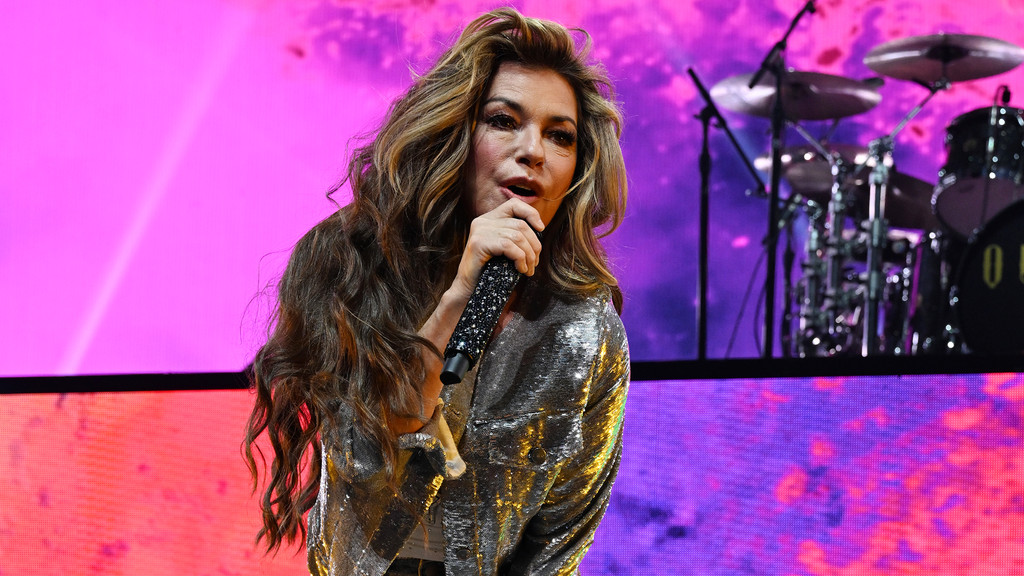 Shania Twain sings into a sparkling microphone on stage, wearing a silver sequined top, with long wavy hair and a vibrant pink and purple screen background.