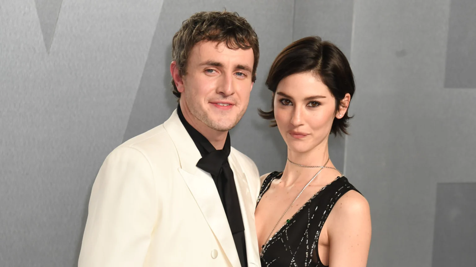 A man in an off-white suit and a woman in a black dress stand side-by-side, looking at the camera against a gray background.