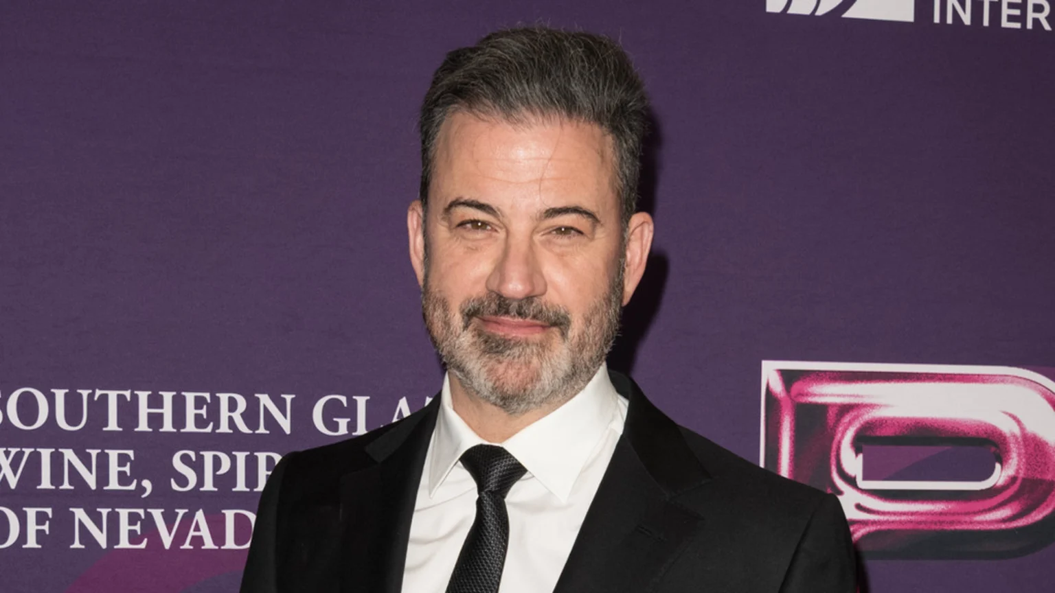 Jimmy Kimmel, a man with graying hair and beard, smiles in a black suit and tie against a purple backdrop with white text and a partially visible pink 'D' logo.