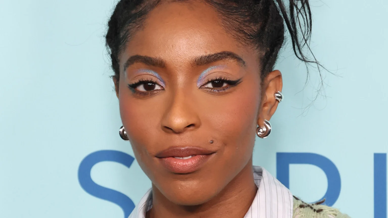 A close-up portrait shows a woman with dark skin, braided hair, shimmery blue eyeshadow, winged eyeliner, and silver spiral earrings, against a light blue background with a partial blue logo.