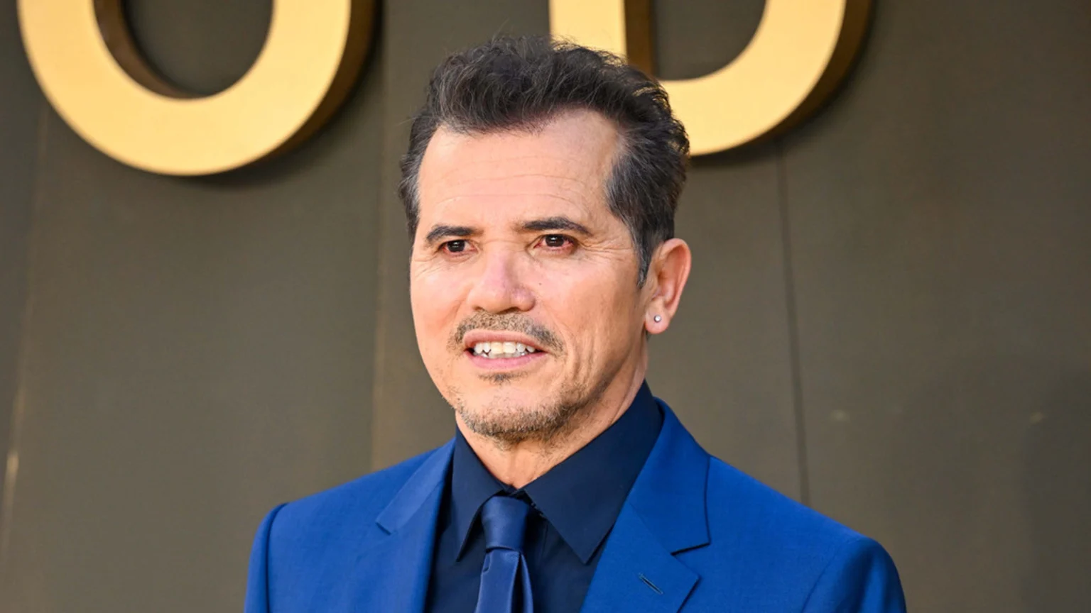 John Leguizamo smiles, wearing a bright blue suit, black shirt, and blue tie, with a mustache, goatee, and a silver stud earring, against a dark background with a golden letter "O".