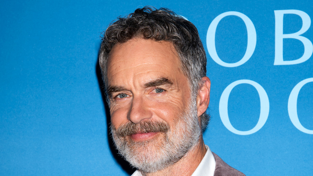 Close-up of actor Murray Bartlett smiling, featuring his grey curly hair, beard, and blue eyes against a blue background with blurred white text.