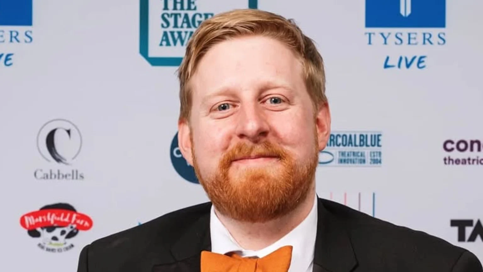 A smiling man with red hair and a red beard, wearing a black suit and orange bow tie, stands in front of a white background with various logos.