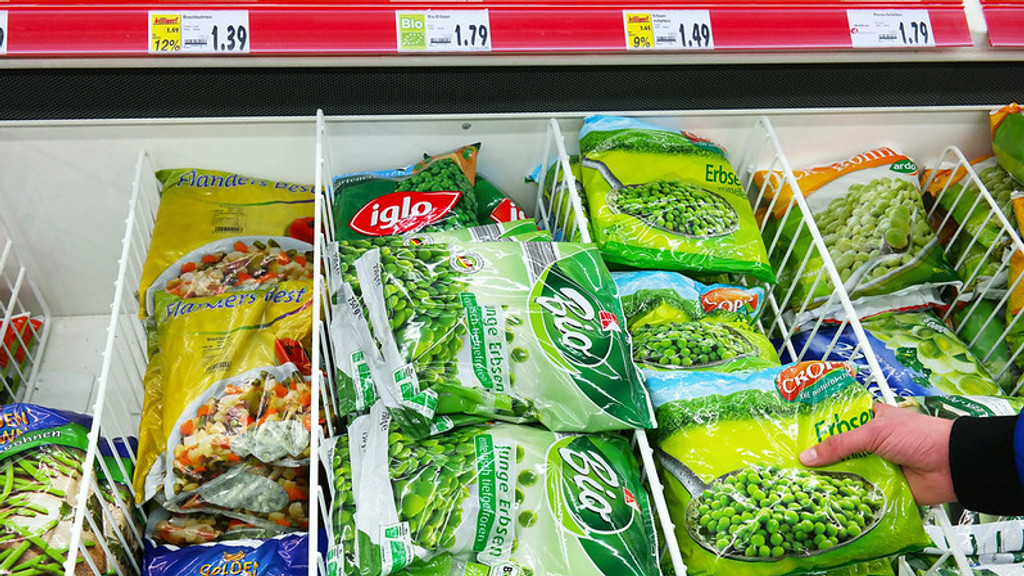 A supermarket freezer aisle displays various bags of frozen vegetables, including peas and mixed vegetables, with price tags above them, and a hand reaching for a bag of peas.