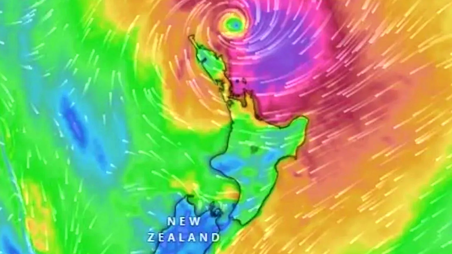 A colorful weather map of New Zealand shows a large, swirling cyclone with an eye centered north of the North Island, indicated by a rainbow of colors and white wind streaks.