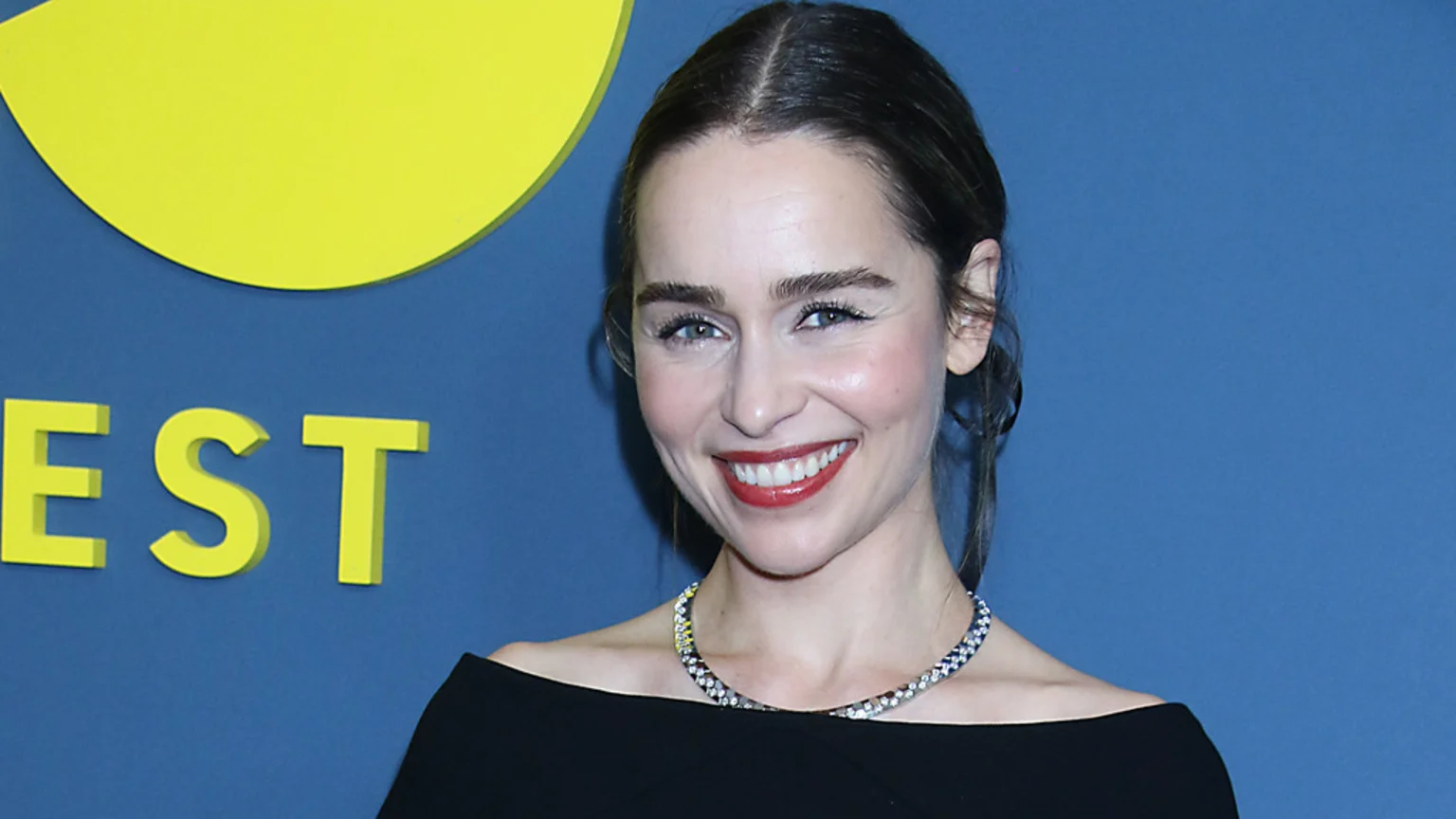 Emilia Clarke smiles at the camera, wearing a black off-the-shoulder top and a sparkling necklace, against a blue background with yellow letters.
