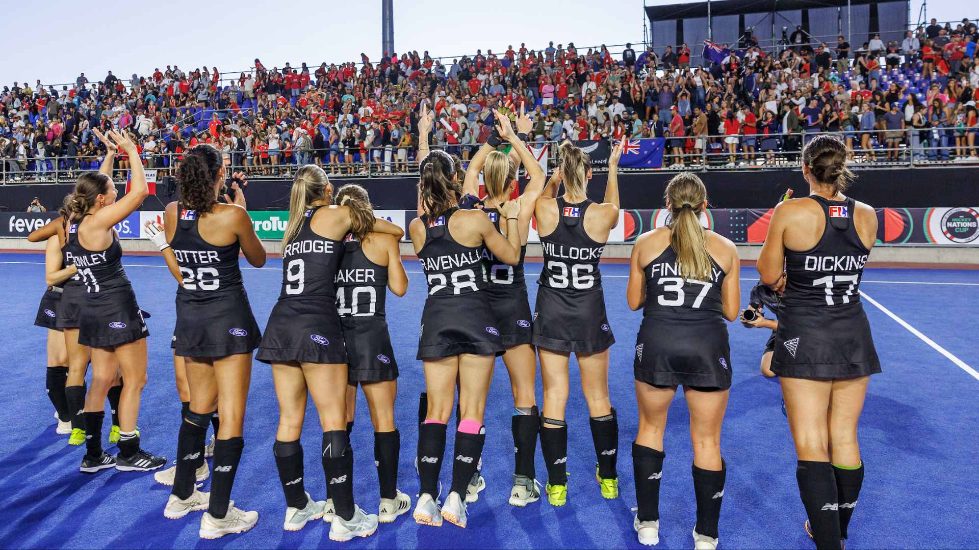 Women's hockey players in black uniforms, seen from behind, raise their arms towards a cheering crowd in a stadium with banners, including one for the Hockey Nations Cup.