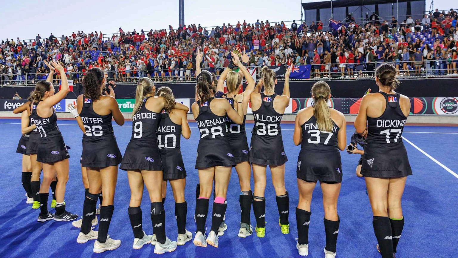 Women's hockey players in black uniforms, seen from behind, raise their arms towards a cheering crowd in a stadium with banners, including one for the Hockey Nations Cup.