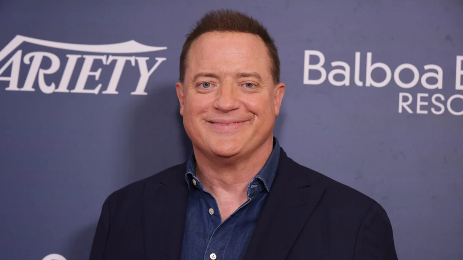 Actor Brendan Fraser smiles, wearing a dark blue suit jacket over a denim shirt, against a purple background with white "Variety" and "Balboa" logos.