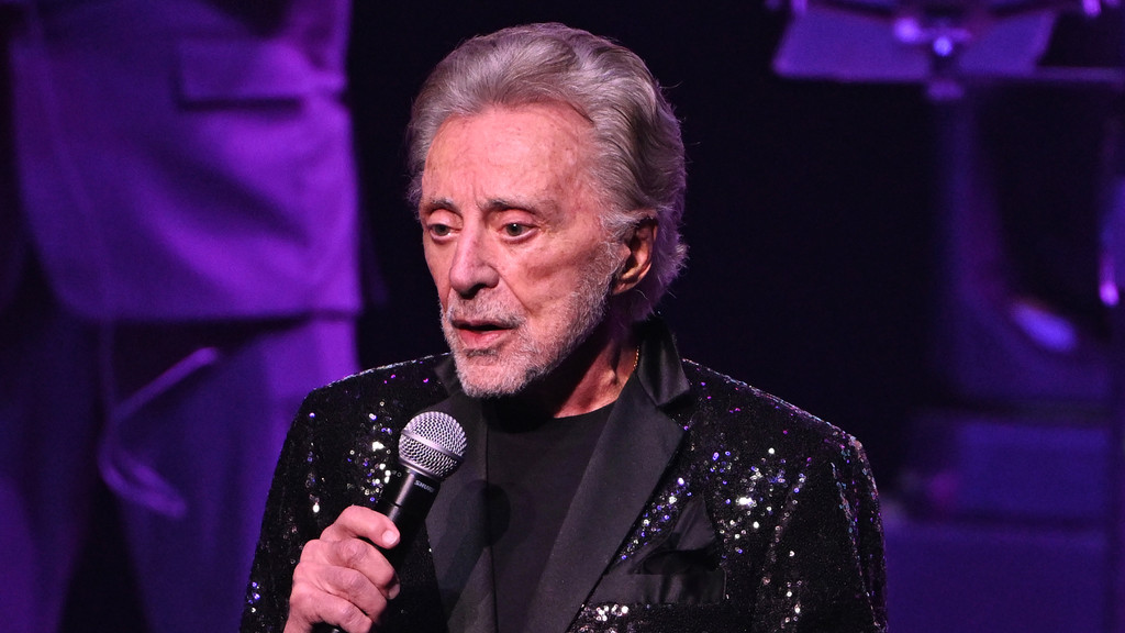A male performer with gray hair and a beard, dressed in a black sequined jacket, holds a microphone on a purple-lit stage.