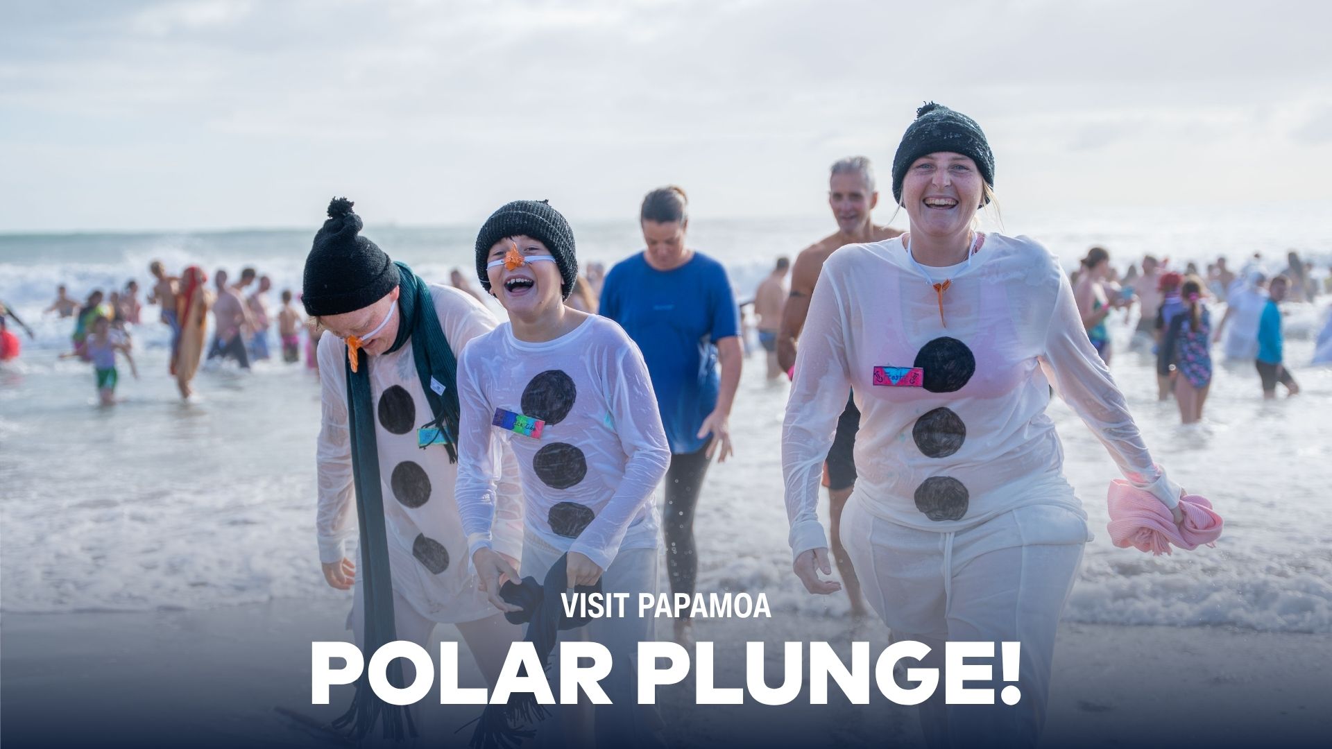 Three smiling people dressed as snowmen emerge from the ocean during a "Polar Plunge" event, with many others in the water in the background.