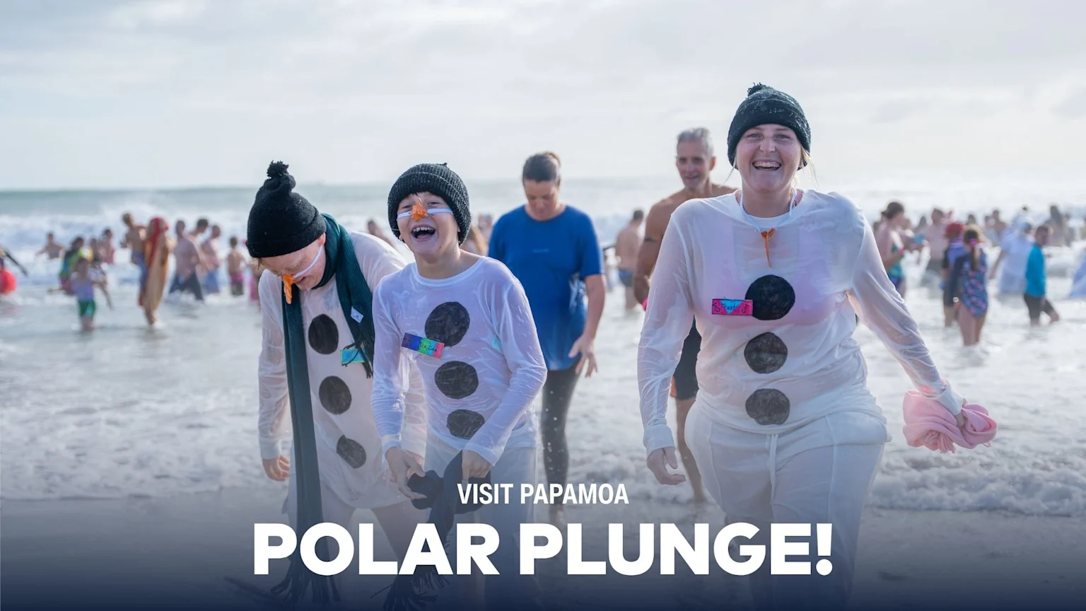 Three smiling people dressed as snowmen emerge from the ocean during a "Polar Plunge" event, with many others in the water in the background.